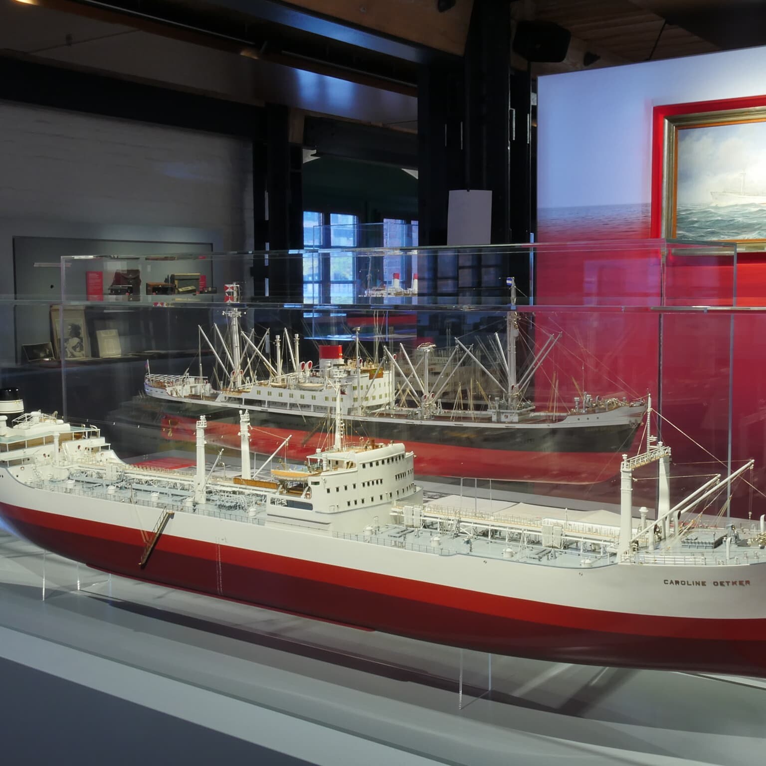 Zwei detaillierte Schiffsmodelle mit rot-weißen Rümpfen in einer Glasvitrine eines Museums, im Hintergrund ein weiteres Modell und ein maritimes Gemälde.