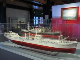 Zwei detaillierte Schiffsmodelle mit rot-weißen Rümpfen in einer Glasvitrine eines Museums, im Hintergrund ein weiteres Modell und ein maritimes Gemälde.