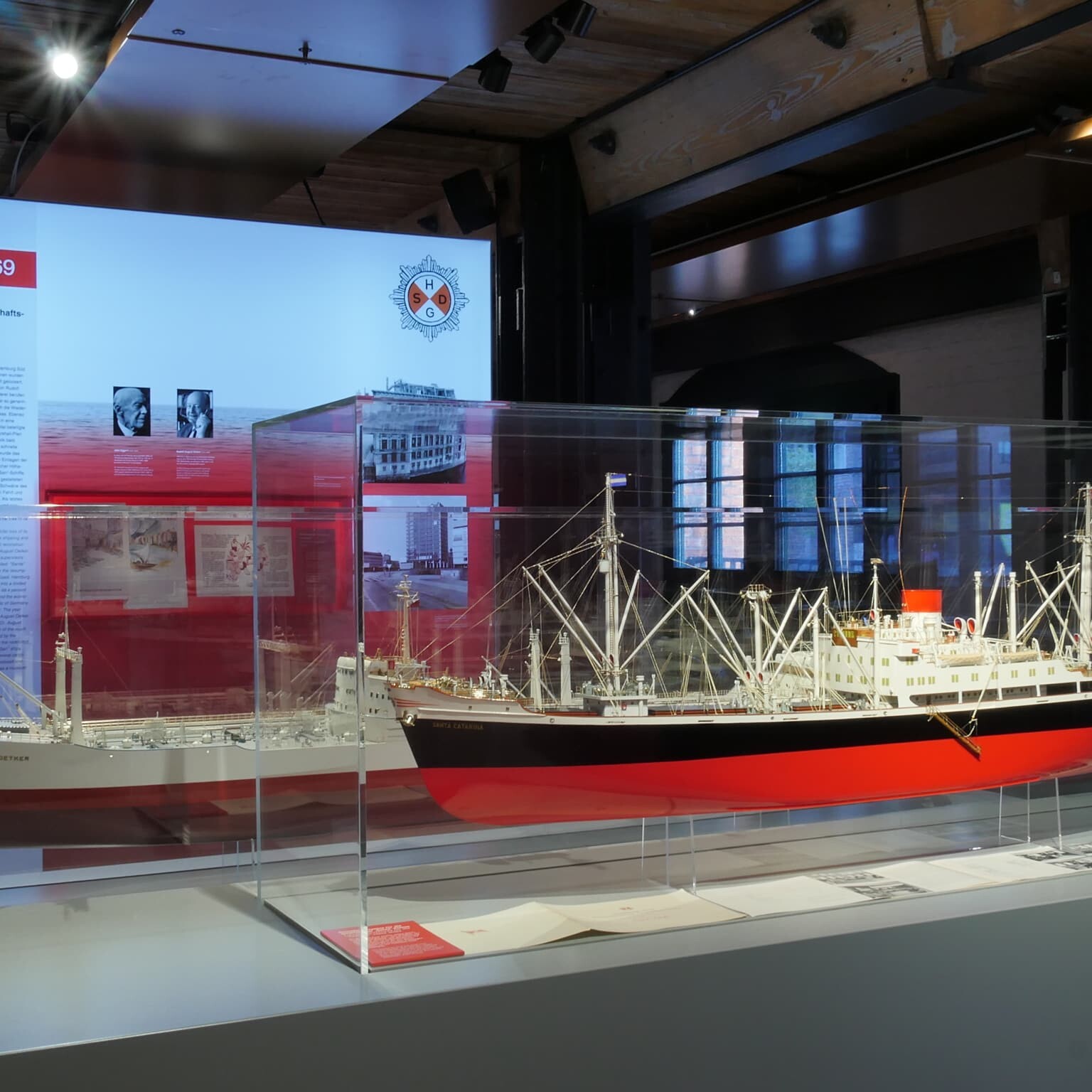 Große Museumsvitrine mit detailliertem rot‑schwarzen Schiffsmodell mit Masten und Takelage, davor Ausstellungstafeln und gedämpfter Deckenbeleuchtung in der Halle.
