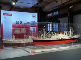 Große Museumsvitrine mit detailliertem rot‑schwarzen Schiffsmodell mit Masten und Takelage, davor Ausstellungstafeln und gedämpfter Deckenbeleuchtung in der Halle.