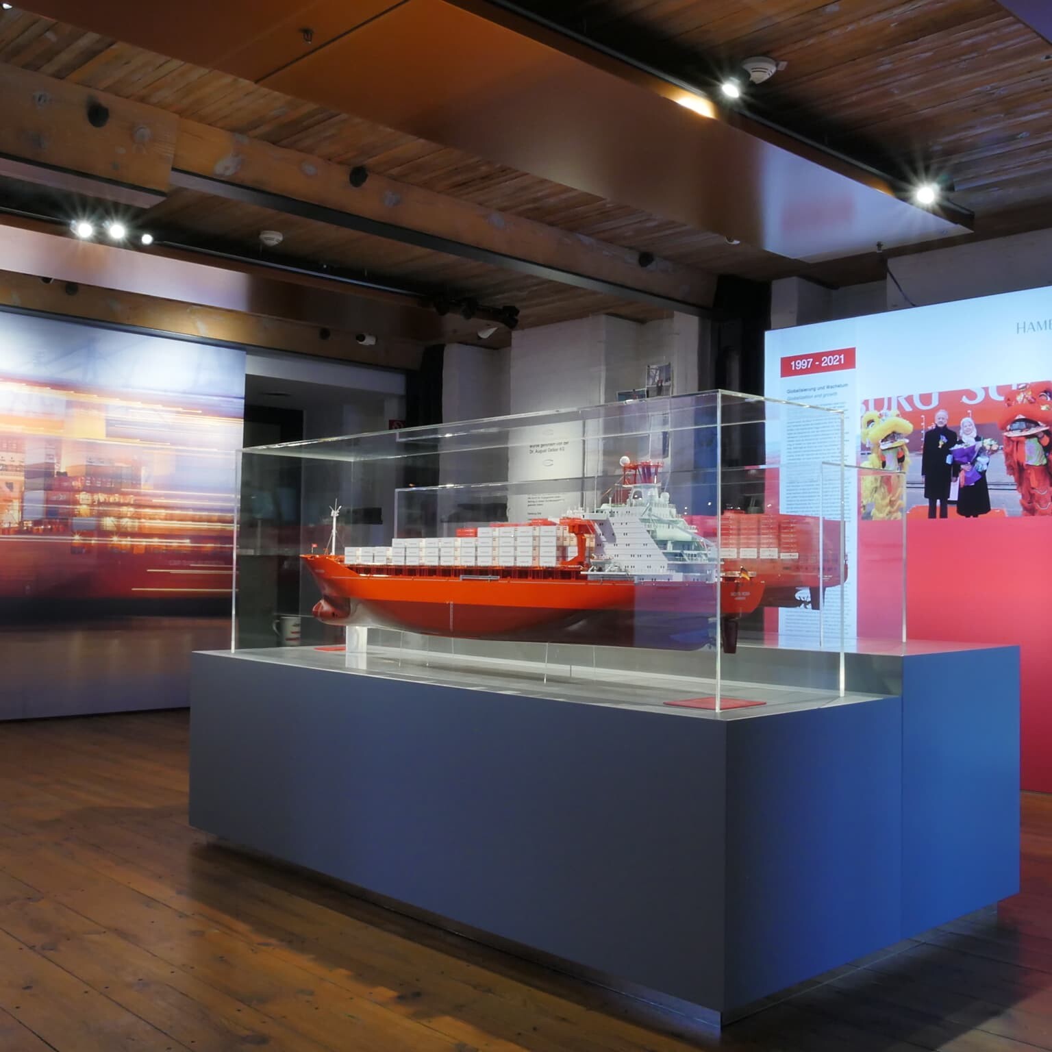 Modell eines rot lackierten Containerschiffs in einer Glasvitrine auf Sockel in einem Museumssaal mit Holzboden, Holzbalkendecke und Hamburg Süd-Displays im Hintergrund.