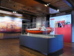 Modell eines rot lackierten Containerschiffs in einer Glasvitrine auf Sockel in einem Museumssaal mit Holzboden, Holzbalkendecke und Hamburg Süd-Displays im Hintergrund.