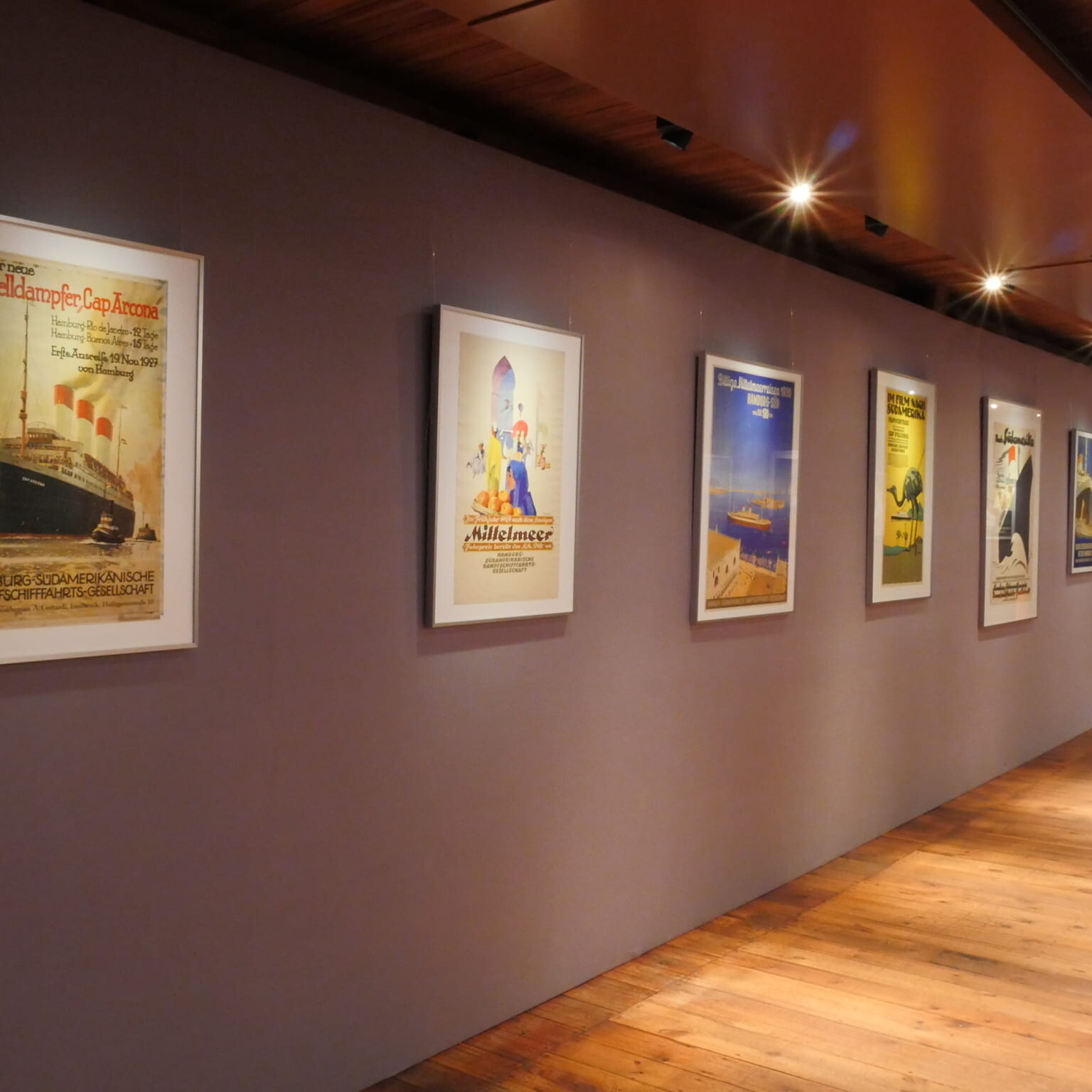 Flur einer Galerie mit Holzboden und Deckenstrahlern, an lilafarbener Wand mehrere gerahmte Vintage-Reiseplakate in Reihen aufgehängt, einheitliche Perspektive.