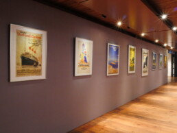 Flur einer Galerie mit Holzboden und Deckenstrahlern, an lilafarbener Wand mehrere gerahmte Vintage-Reiseplakate in Reihen aufgehängt, einheitliche Perspektive.