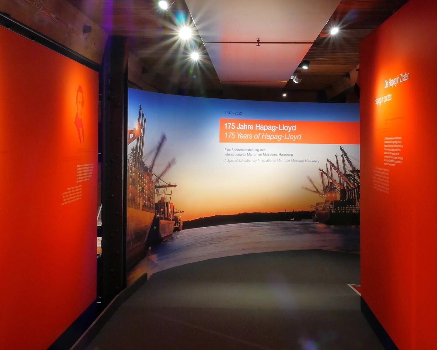Ausstellungsflur mit orangen W&auml;nden und Portraits, am Ende Tafel '175 Jahre Hapag‑Lloyd' vor Containerschiffen und Hafenkr&auml;nen im Abendlicht.