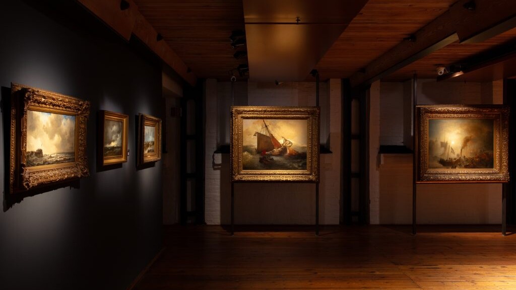 Dimmer Museumsraum mit warmem Holzboden, mehreren goldgerahmten Seestücken an dunklen Wänden und einem beleuchteten Gemälde eines Segelboots.