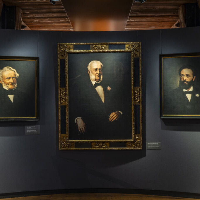 Drei gerahmte Ölporträts älterer Herren in dunkler Kleidung und Fliege an einer grauen Museumswand, mit einem größeren, reich verzierten Goldrahmen in der Mitte und zwei kleineren schwarzen Rahmen links und rechts.