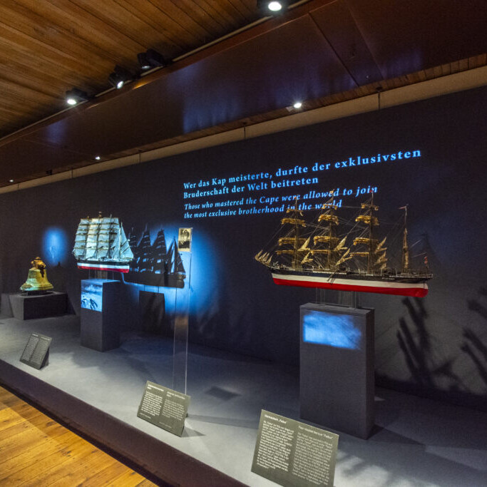 Museumsvitrine mit mehreren beleuchteten Segelschiffsmodellen auf Podesten vor einer dunklen Wand, auf der deutscher und englischer Text zur Kapitelsbruderschaft projiziert ist; links sichtbar der Schriftzug '200 Jahre F. Laeisz' und die Holzdecke sowie der Parkettboden rahmen die Szene.