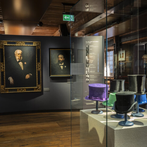 Museumsausstellung: links drei formelle Porträtgemälde an einer dunklen Wand, rechts eine Glasvitrine mit mehreren bunten Zylinderhüten auf Podesten, im Raum sichtbare Holzdecke und -boden sowie gedämpfte Beleuchtung.