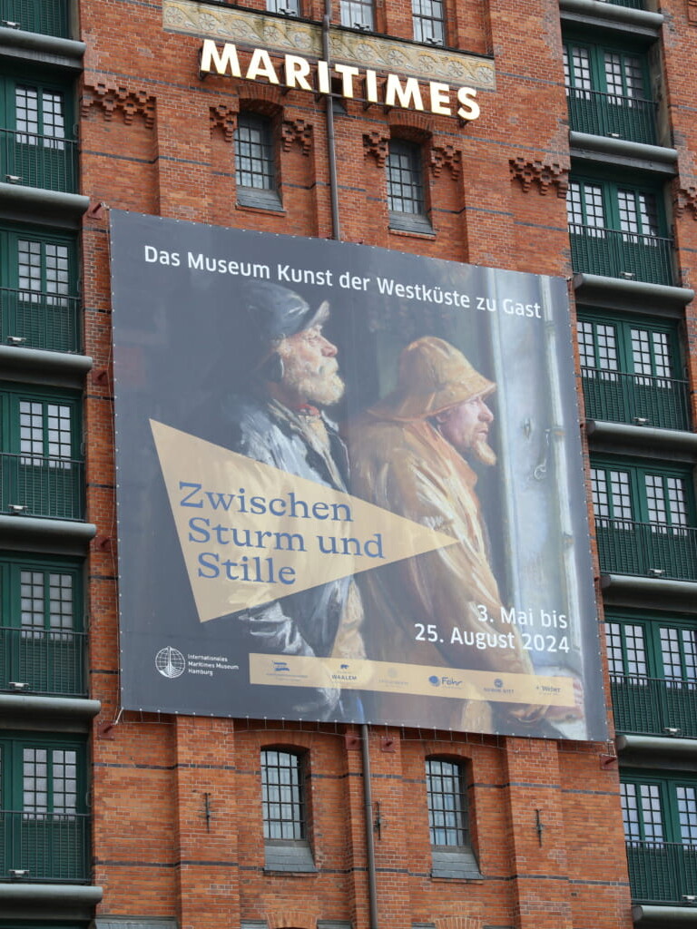 Rote Backsteinfassade mit leuchtendem Schriftzug MARITIMES und großem Banner 'Zwischen Sturm und Stille' mit zwei wettergegerbten Fischern in Regenmänteln.