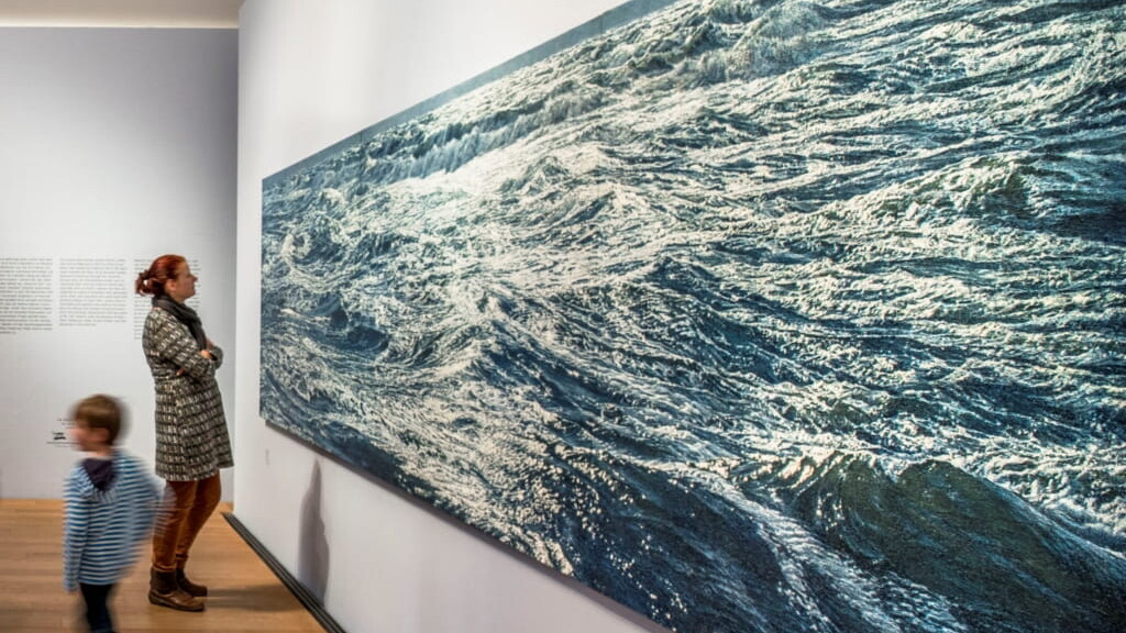 Frau betrachtet in einer Galerie ein großformatiges, wellenbewegtes Meeresbild an weißer Wand, im Vordergrund läuft ein verschwommener Junge vorbei.