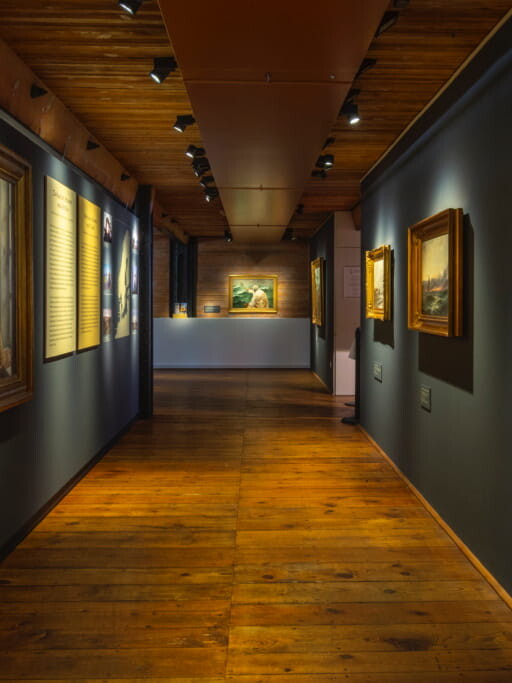 Schmaler, stimmungsvoll beleuchteter Museumsflur mit warmem Holzfußboden und -decke, graue Wände mit vergoldeten Gemälderahmen und Blick zur Empfangstheke.