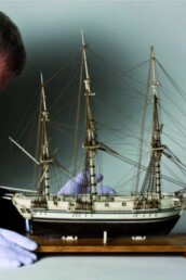 Älterer Mann mit Handschuhen und Brille richtet behutsam ein detailreiches Segelschiffsmodell mit Takelage auf einem Holzsockel vor neutralem Hintergrund.