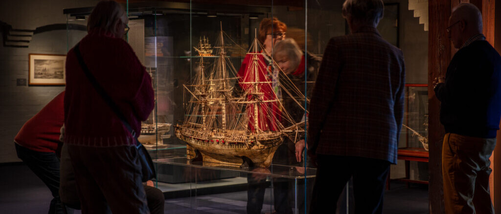 Mehrere Besucher betrachten ein detailliertes Schiffsmodell mit roten Segeln in einer Glasvitrine in einem Museum.