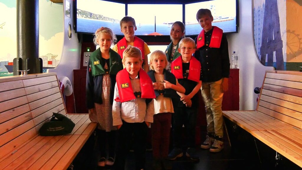 Sieben Kinder mit roten und grünen Rettungswesten stehen in einem Schiffssimulator vor drei Bildschirmen, umgeben von Holzbänken und maritimen Wandbildern.