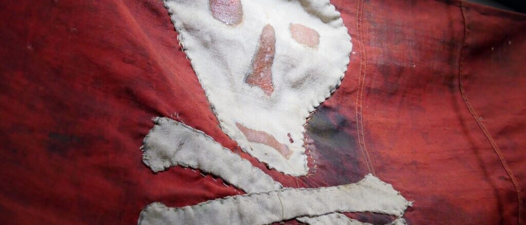 Rotes, abgenutztes Stofftuch mit grob aufgenähtem weißen Totenkopf und gekreuzten Knochen, fransige Ränder sichtbar.