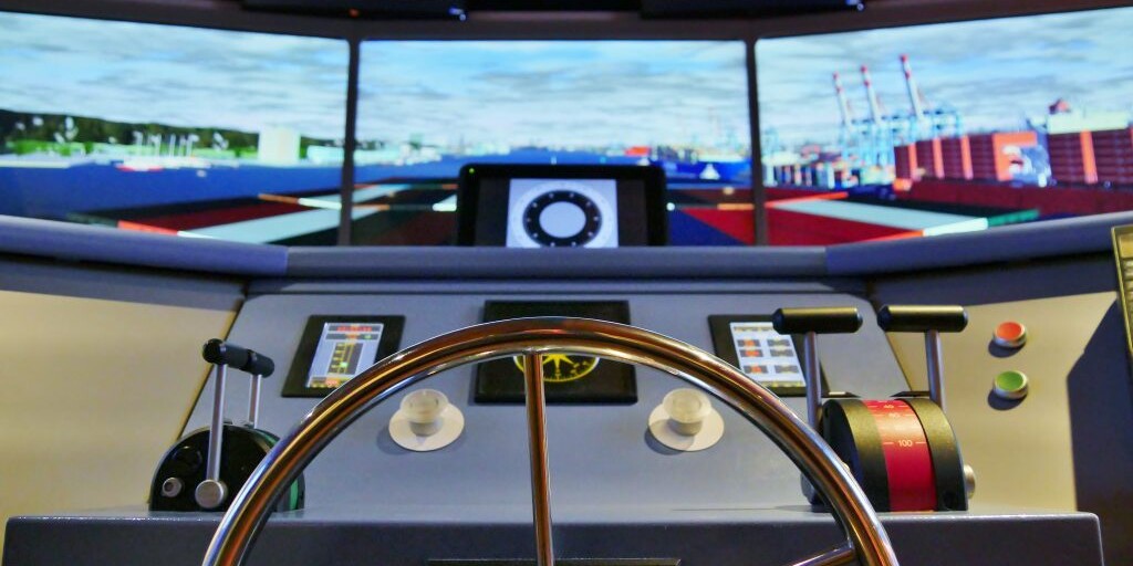 Blick über ein poliertes Metallsteuerrad auf die Brücke eines Schiffs-Simulators mit Schubhebeln, Knöpfen und mehreren Bildschirmen, die einen Hafen mit Containern zeigen.