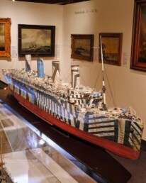 Großes Schiffsmodell in einem Museum mit auffälliger Dazzle-Tarnung aus schwarzen und weißen geometrischen Streifen, rotem Rumpf und drei Schornsteinen.
