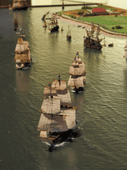 Diorama mit mehreren historischen Dreimast-Segelschiffen in Formation auf ruhigem Wasser, mit Booten und einer grünen Küstenlandschaft im Hintergrund.