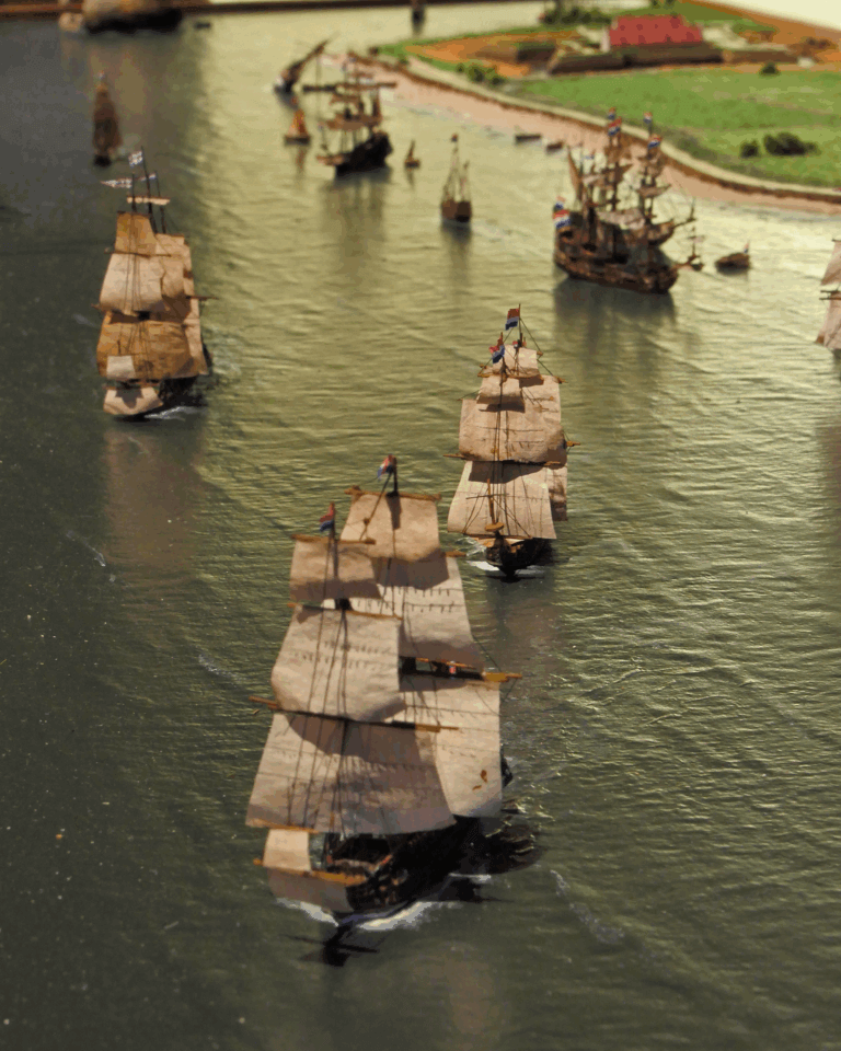 Diorama mit mehreren historischen Dreimast-Segelschiffen in Formation auf ruhigem Wasser, mit Booten und einer grünen Küstenlandschaft im Hintergrund.