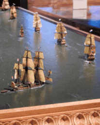 Mehrere detailreiche Modellsegelschiffe mit beige-gelben Segeln auf einem Wasser-Diorama, im Vordergrund ein größeres Dreimastschiff und im Bildrand ein geschnitzter Holzrahmen.