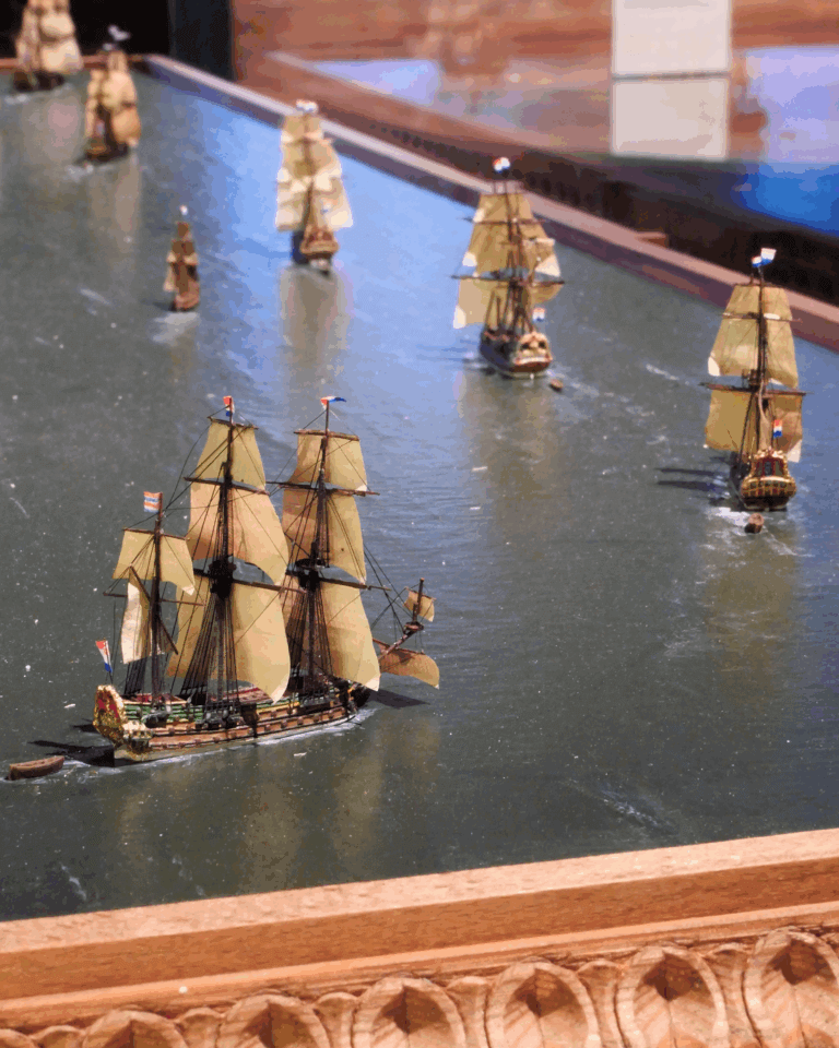 Mehrere detailreiche Modellsegelschiffe mit beige-gelben Segeln auf einem Wasser-Diorama, im Vordergrund ein größeres Dreimastschiff und im Bildrand ein geschnitzter Holzrahmen.