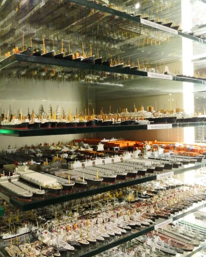Glasregale voller hunderter kleiner Schiffsmodelle verschiedener Typen und Größen, eng gereiht mit Masten, Containern und Lichtreflexen hinter Glasscheiben.