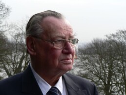 &Auml;lterer Mann im Anzug und Krawatte mit Brille, seitliches Portr&auml;t vor kahlen B&auml;umen und einem Gew&auml;sser im Hintergrund