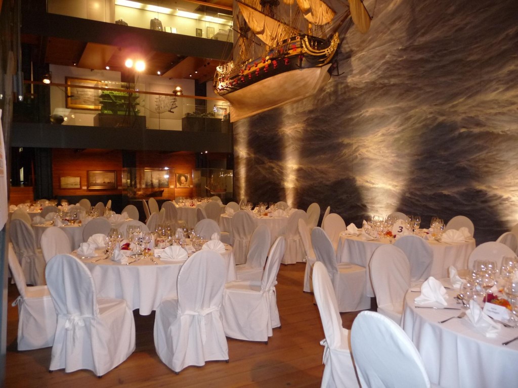 Eleganter Festsaal mit rundgedeckten Tischen und weißen Stuhlhussen, Gläsern und Servietten, darüber ein hängendes Schiffsmodell vor einer Wand mit Wellenmotiv.