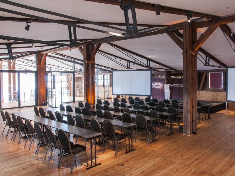 Konferenzraum in Holzbalkenhalle mit Reihen von Tischen und Stühlen, zwei Projektionsleinwänden und Holzfußboden.