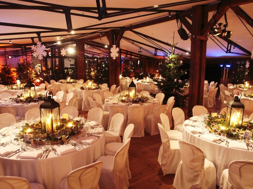 Weihnachtlich dekorierter Festsaal mit runden Tischen, weißen Stuhlhussen, Laternen als Mittelstücke, Girlanden und Tannenbäumen.