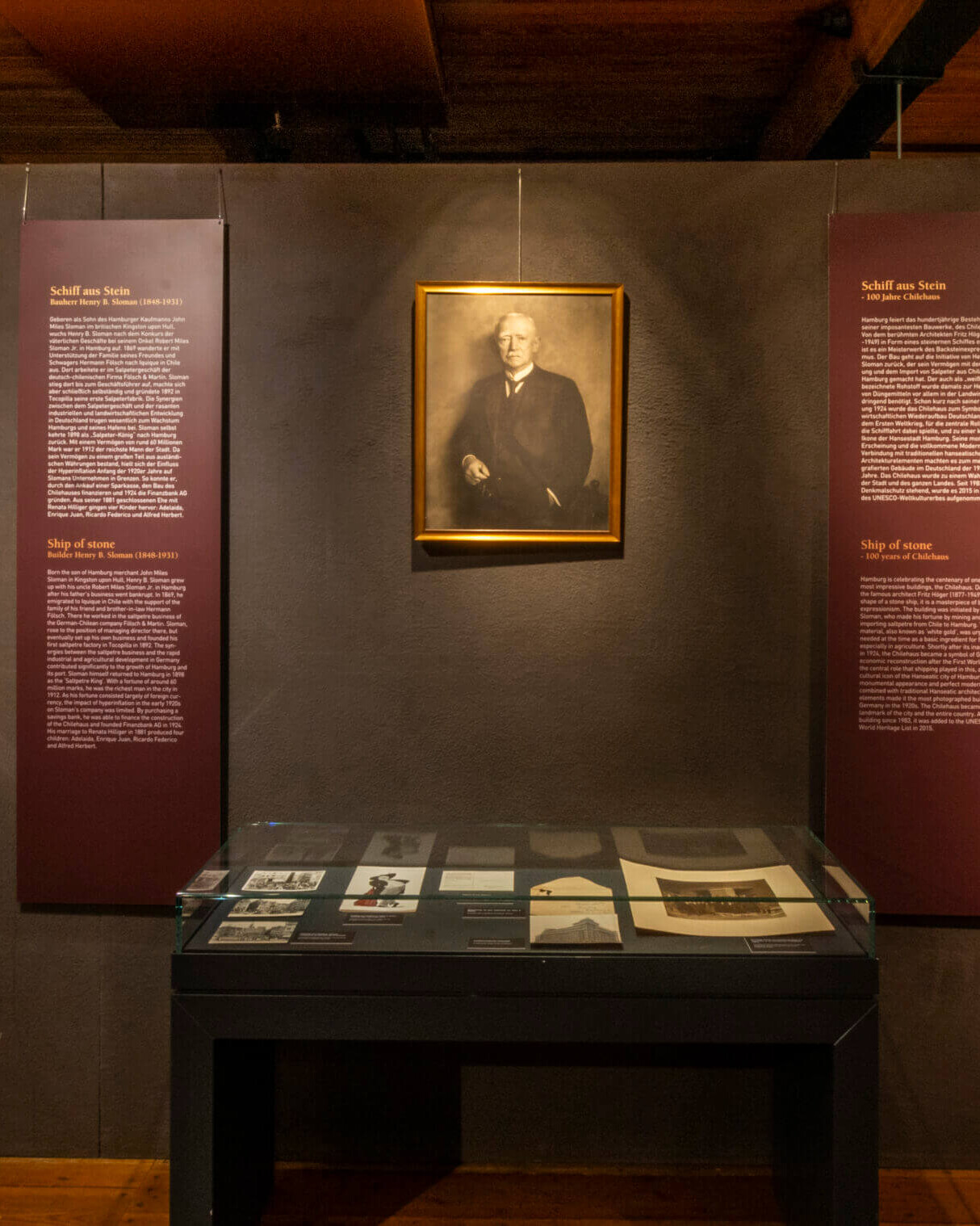 Museumsausstellung in einem holzgetäfelten Raum: mittig an einer dunklen Wand ein gerahmtes Porträt, darunter eine Glasvitrine mit historischen Dokumenten, links eine Tafel mit neun alten Fotografien und erläuterndem Text, rechts weitere Informationstafeln und ein auf einem Staffelei-ähnlichen Gestell stehendes großes Foto.