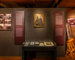 Museumsausstellung in einem holzgetäfelten Raum: mittig an einer dunklen Wand ein gerahmtes Porträt, darunter eine Glasvitrine mit historischen Dokumenten, links eine Tafel mit neun alten Fotografien und erläuterndem Text, rechts weitere Informationstafeln und ein auf einem Staffelei-ähnlichen Gestell stehendes großes Foto.
