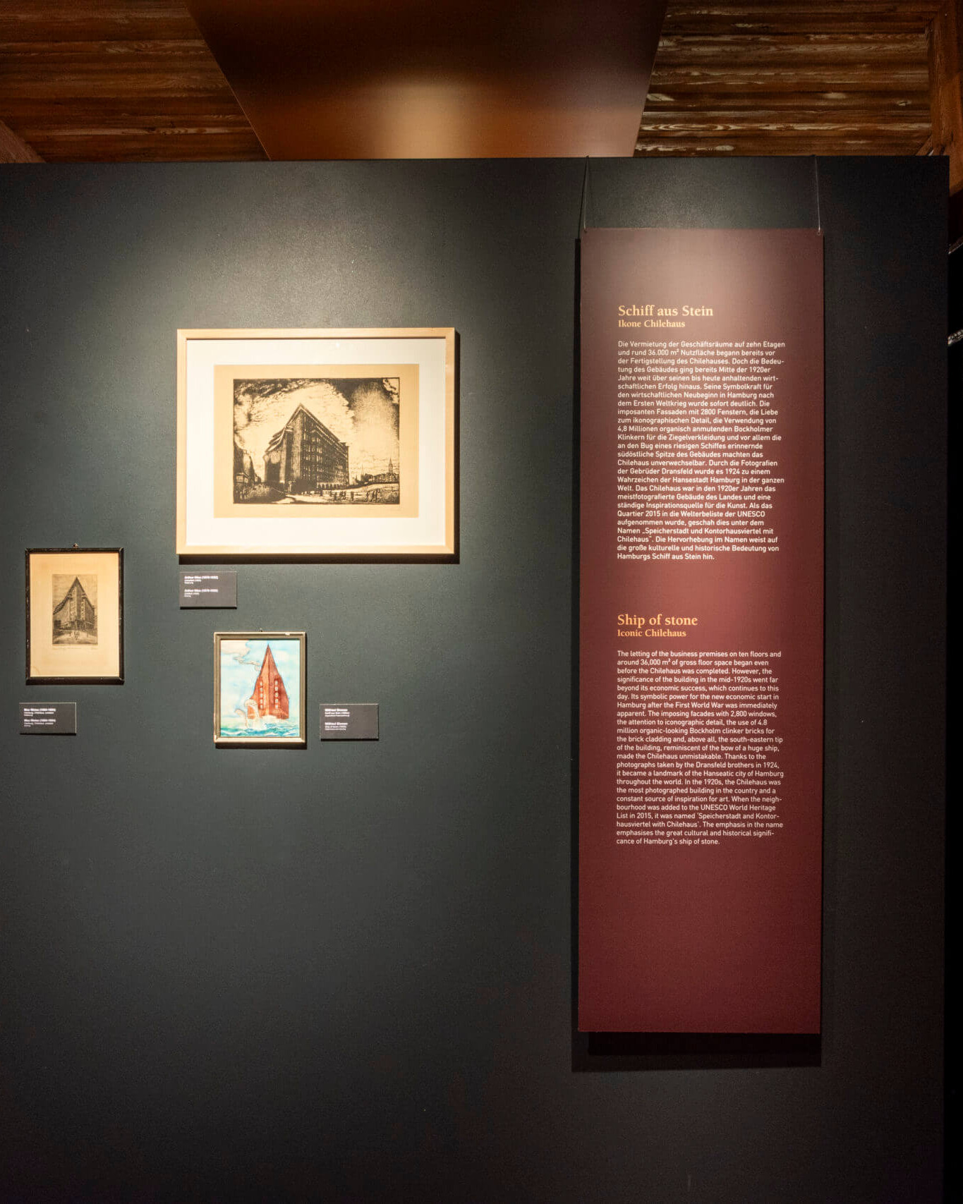 Ausstellungswand in einem Museum mit drei gerahmten Grafiken – einem großen schwarz-weißen Druck oben und zwei kleineren darunter (eins farbig mit einem roten Schiff) – neben einer hohen dunkelroten Informationstafel mit der Überschrift „Schiff aus Stein“, links und rechts von sichtbaren schwarzen Stahlträgern und Holzverkleidungen eingerahmt.