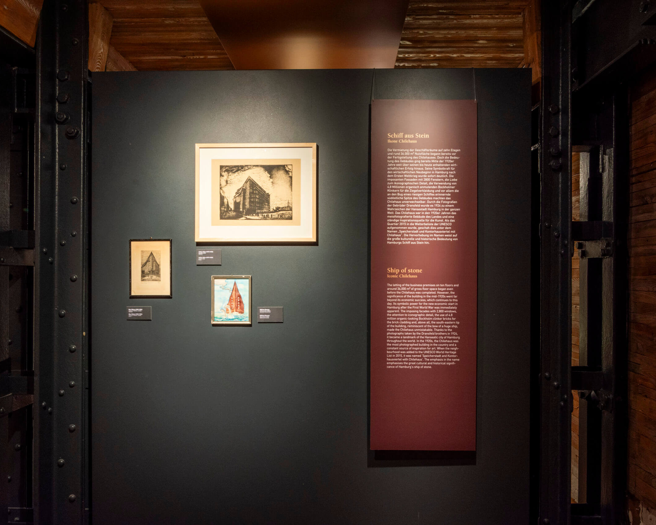 Ausstellungswand in einem Museum mit drei gerahmten Grafiken – einem großen schwarz-weißen Druck oben und zwei kleineren darunter (eins farbig mit einem roten Schiff) – neben einer hohen dunkelroten Informationstafel mit der Überschrift „Schiff aus Stein“, links und rechts von sichtbaren schwarzen Stahlträgern und Holzverkleidungen eingerahmt.