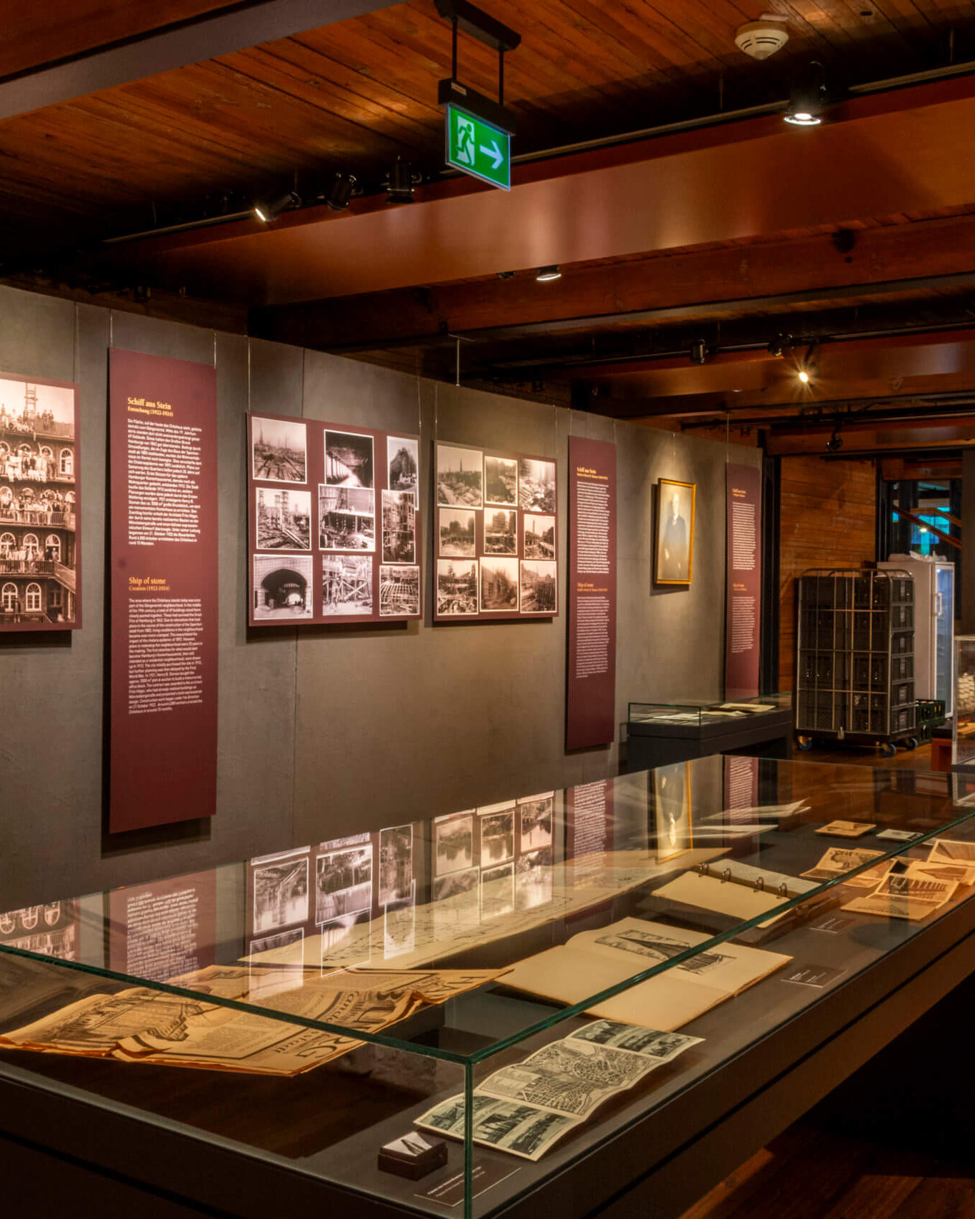Innenraum eines Museums mit warmem Holzdekor, mehreren Glasvitrinen mit historischen Dokumenten und Drucken, Wandtafeln mit sepiafarbigen Fotografien sowie einem gerahmten Gemälde eines Segelschiffs unter sichtbaren Deckenbalken und gedämpfter Beleuchtung.