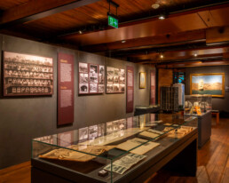Innenraum eines Museums mit warmem Holzdekor, mehreren Glasvitrinen mit historischen Dokumenten und Drucken, Wandtafeln mit sepiafarbigen Fotografien sowie einem gerahmten Gemälde eines Segelschiffs unter sichtbaren Deckenbalken und gedämpfter Beleuchtung.