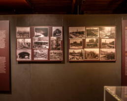 Museumswand mit drei dunkelroten Informationstafeln – zwei schmale Textfelder links und rechts und in der Mitte zwei große Tafeln mit mehreren historischen Sepia-/Schwarzweiß-Fotografien und erklärendem Text über das Bauprojekt „Schiff aus Stein“, rechts unten eine Glasvitrine sichtbar.