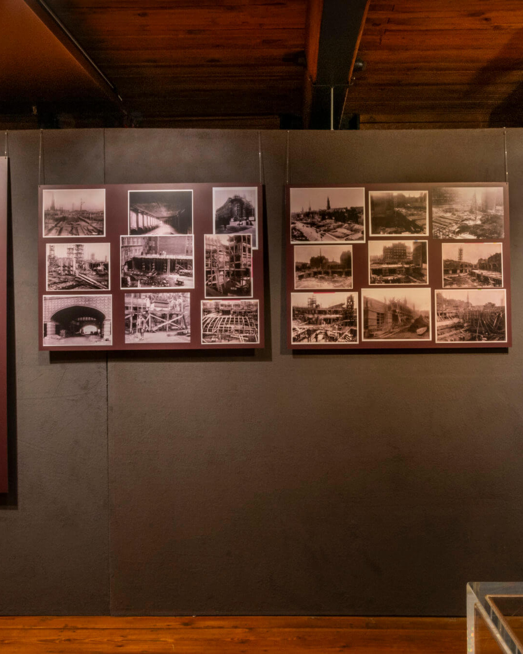 Museumsausstellungswand mit mehreren gerahmten sepiafarbenen historischen Fotografien und zwei hohen roten Informationstafeln in der Mitte, rechts ein gerahmtes Porträt eines Mannes sowie sichtbare Holzdecke oben und Holzfußboden unten.