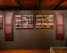 Museumsausstellungswand mit mehreren gerahmten sepiafarbenen historischen Fotografien und zwei hohen roten Informationstafeln in der Mitte, rechts ein gerahmtes Porträt eines Mannes sowie sichtbare Holzdecke oben und Holzfußboden unten.