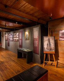 Warm beleuchteter Museumsraum mit Holzfußboden und sichtbaren Deckenbalken, Ausstellungswänden mit historischen Fotografien und Texttafeln, mehreren Glasvitrinen in der Mitte und einer Staffelei mit einem Poster zum Thema Schiff/Chilehaus.