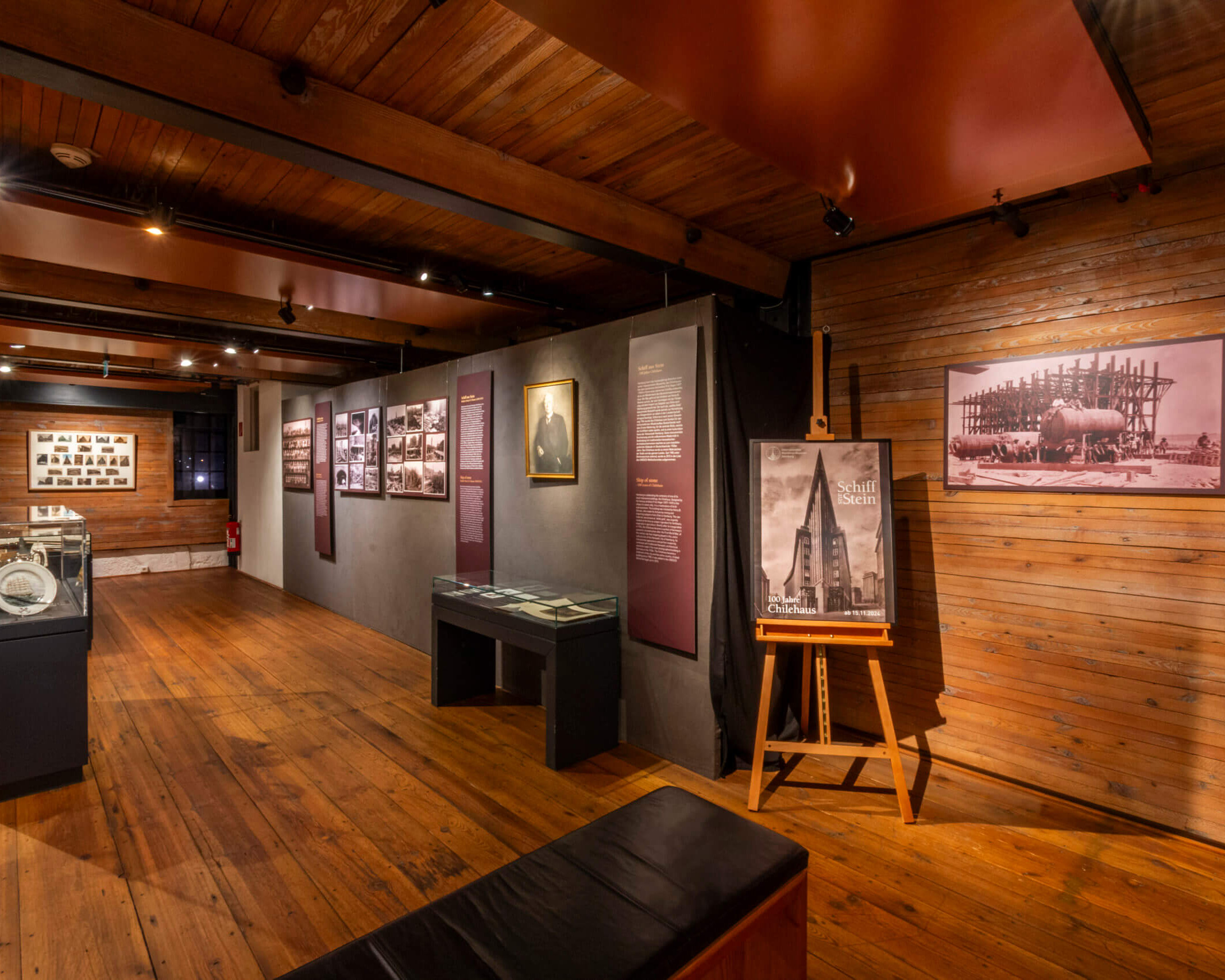Warm beleuchteter Museumsraum mit Holzfußboden und sichtbaren Deckenbalken, Ausstellungswänden mit historischen Fotografien und Texttafeln, mehreren Glasvitrinen in der Mitte und einer Staffelei mit einem Poster zum Thema Schiff/Chilehaus.
