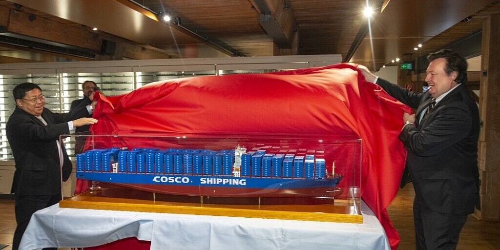Zwei M&auml;nner l&uuml;ften ein gro&szlig;es rotes Tuch und enth&uuml;llen auf einem wei&szlig;en Tisch ein im Glaskasten ausgestelltes Modell-Containerfrachtschiff mit blauen Containern und der Aufschrift COSCO SHIPPING in einem holzverkleideten Innenraum.
