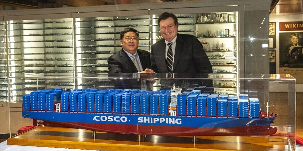 Zwei M&auml;nner in Anz&uuml;gen sch&uuml;tteln l&auml;chelnd die Hand hinter einer gro&szlig;en blau-roten Modellvitrine des Containerschiffs mit der Aufschrift COSCO SHIPPING; im Hintergrund sind beleuchtete Vitrinen mit zahlreichen Schiffsmodellen zu sehen.
