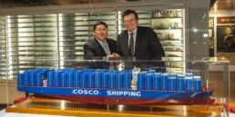 Zwei M&auml;nner in Anz&uuml;gen sch&uuml;tteln l&auml;chelnd die Hand hinter einer gro&szlig;en blau-roten Modellvitrine des Containerschiffs mit der Aufschrift COSCO SHIPPING; im Hintergrund sind beleuchtete Vitrinen mit zahlreichen Schiffsmodellen zu sehen.