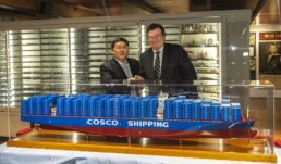 Zwei Männer in Anzügen schütteln lächelnd die Hand hinter einer großen blau-roten Modellvitrine des Containerschiffs mit der Aufschrift COSCO SHIPPING; im Hintergrund sind beleuchtete Vitrinen mit zahlreichen Schiffsmodellen zu sehen.