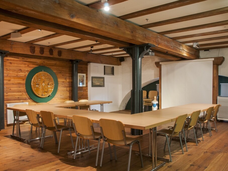 Konferenzraum mit U-förmigen Holztischen und Stühlen, sichtbaren Holzbalken, Holzverkleidung an der Wand und Projektionsleinwand.