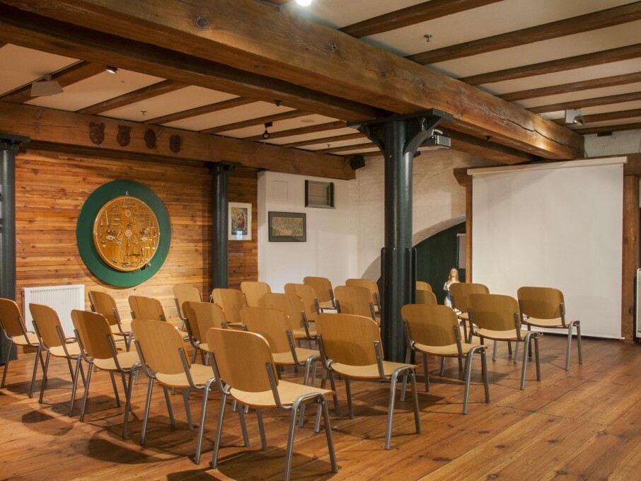 Seminarraum mit Holzboden, mehreren Reihen einfacher Holzstühle, sichtbaren Deckenbalken, Leinwand und rundem Wandrelief.