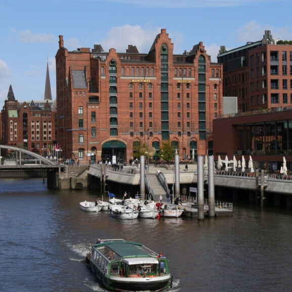 Ausflugsboot fährt auf einem Hafenkanal vor rotem Backsteingebäude mit der Aufschrift „Maritimes Museum“, Uferpromenade mit Stegen und kleineren Booten.