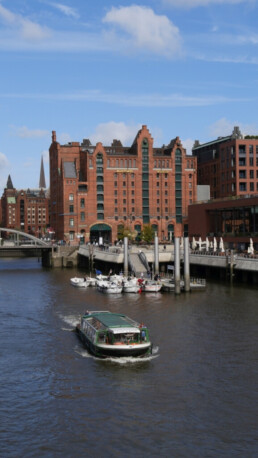 Grünes Passagierschiff fährt in einem Hafenkanal unter blauem Himmel; im Hintergrund ein großes rotes Backsteingebäude mit Giebeln, Schriftzug „Maritimes Museum“, Anleger und Yachten.